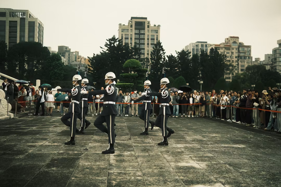 Four honour guards mid-stride with legs raised in a slow high step, crowd lined up along a red rope