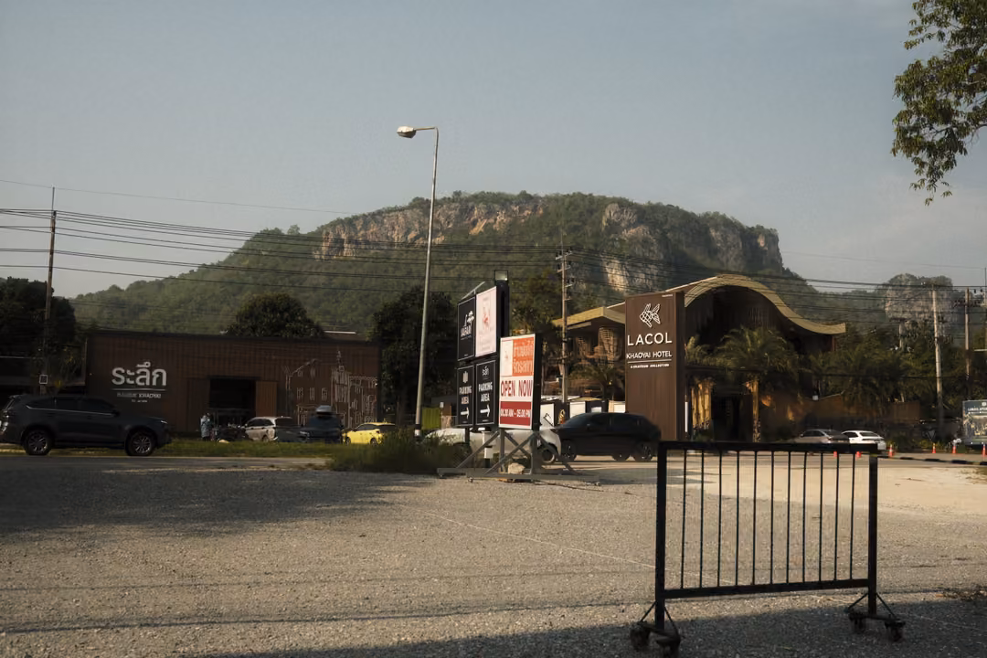 A gravel car park with a metal barrier, low buildings and signs, a craggy limestone hill behind