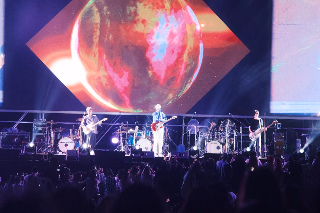 Band performing below a screen showing a glowing red and orange sphere on a diamond background, purple stage light.