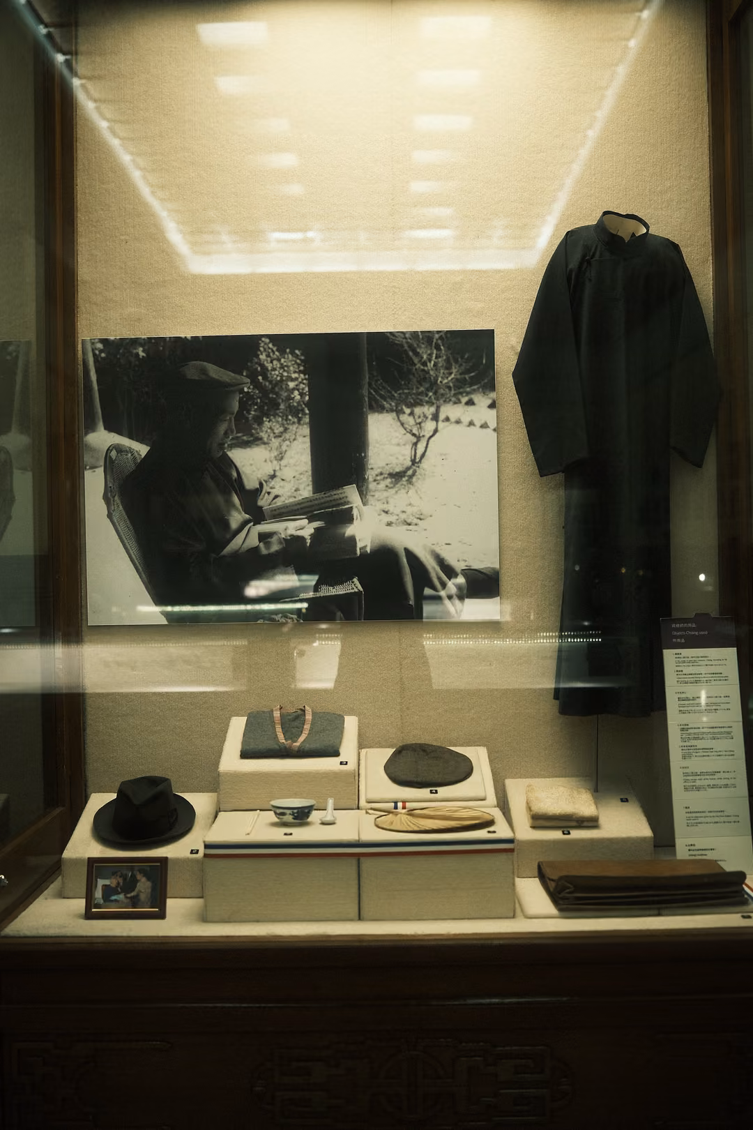 Glass display case with a dark robe, a black-and-white photograph of a seated man reading, a hat and small objects
