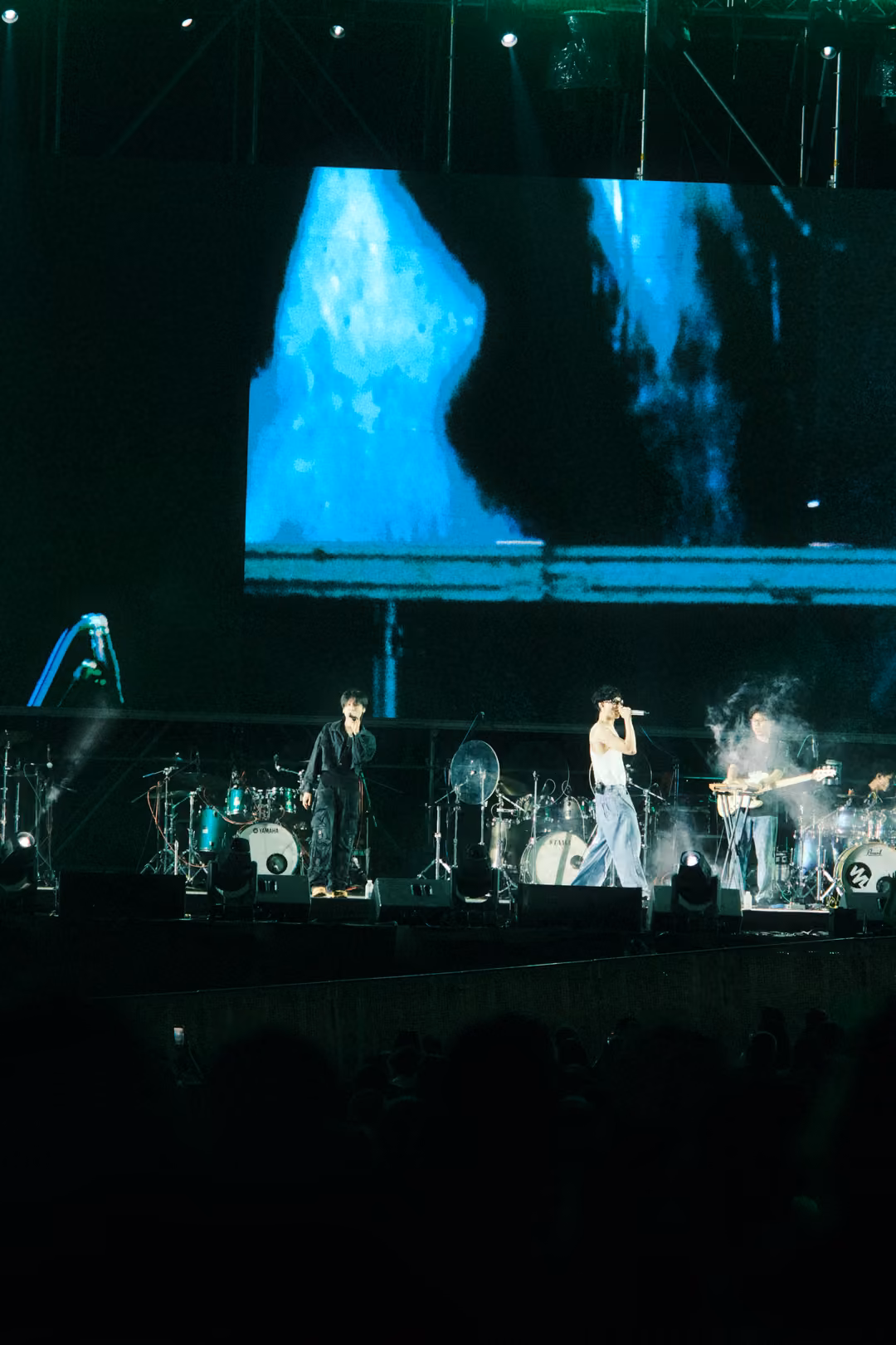 Stage at night with a screen showing blue streaked shapes, the band performing amid swirling smoke.