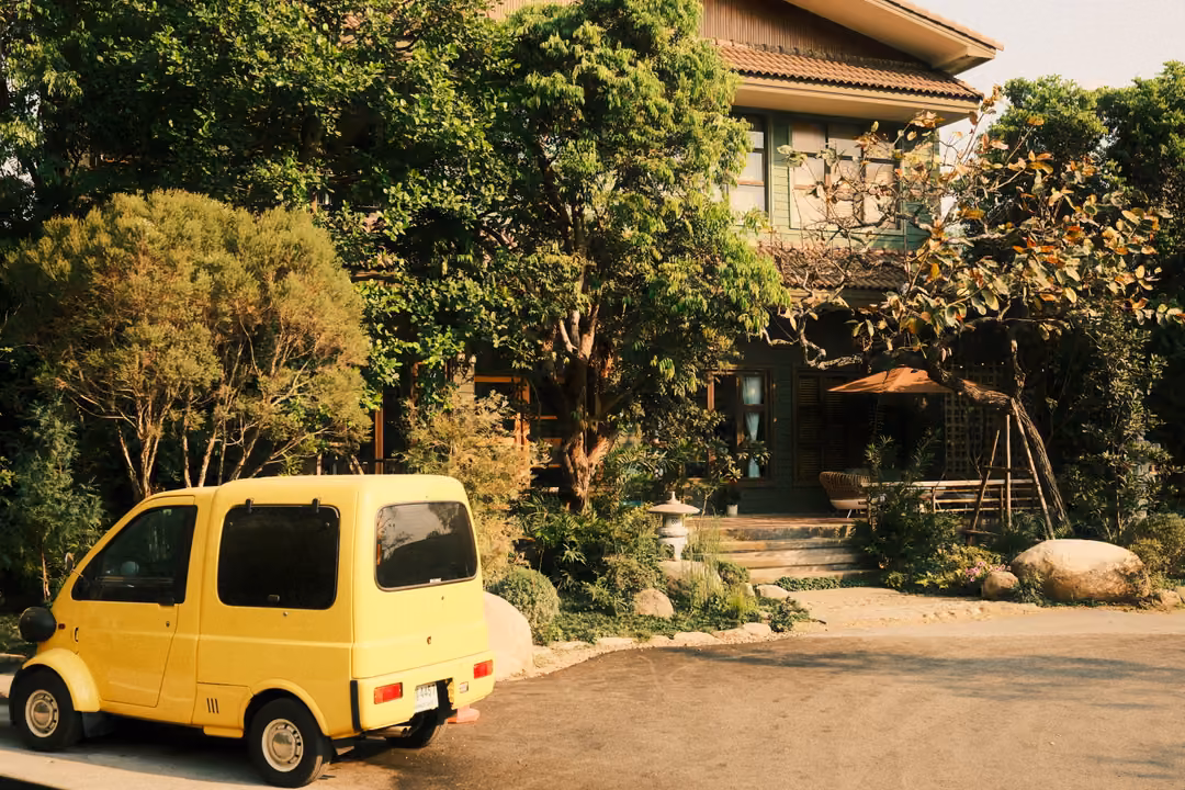 Small boxy yellow car parked in front of a wooden house among green trees, shrubs and boulders