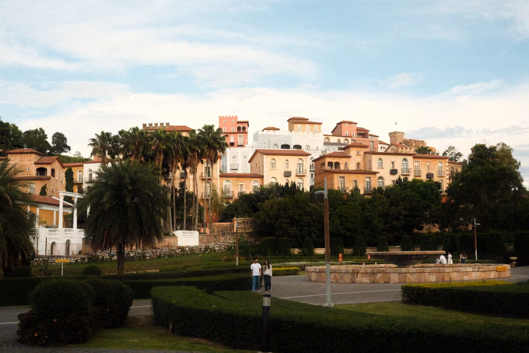 Garden of clipped hedges and palms with a shallow fountain basin and two people standing before a hillside village