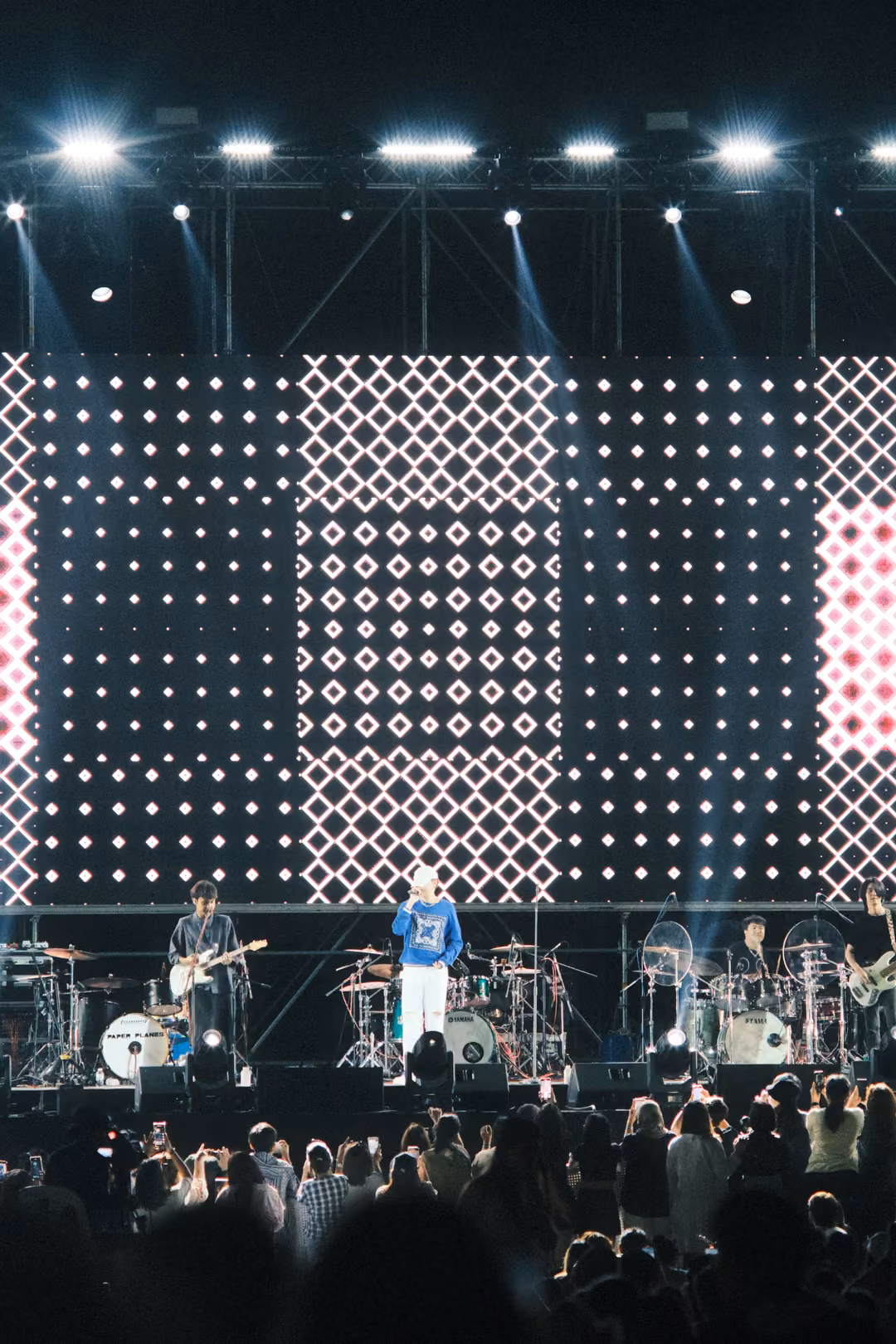 Vertical view of a stage with a screen of white diamond patterns and a pink panel, crowd heads lit below.