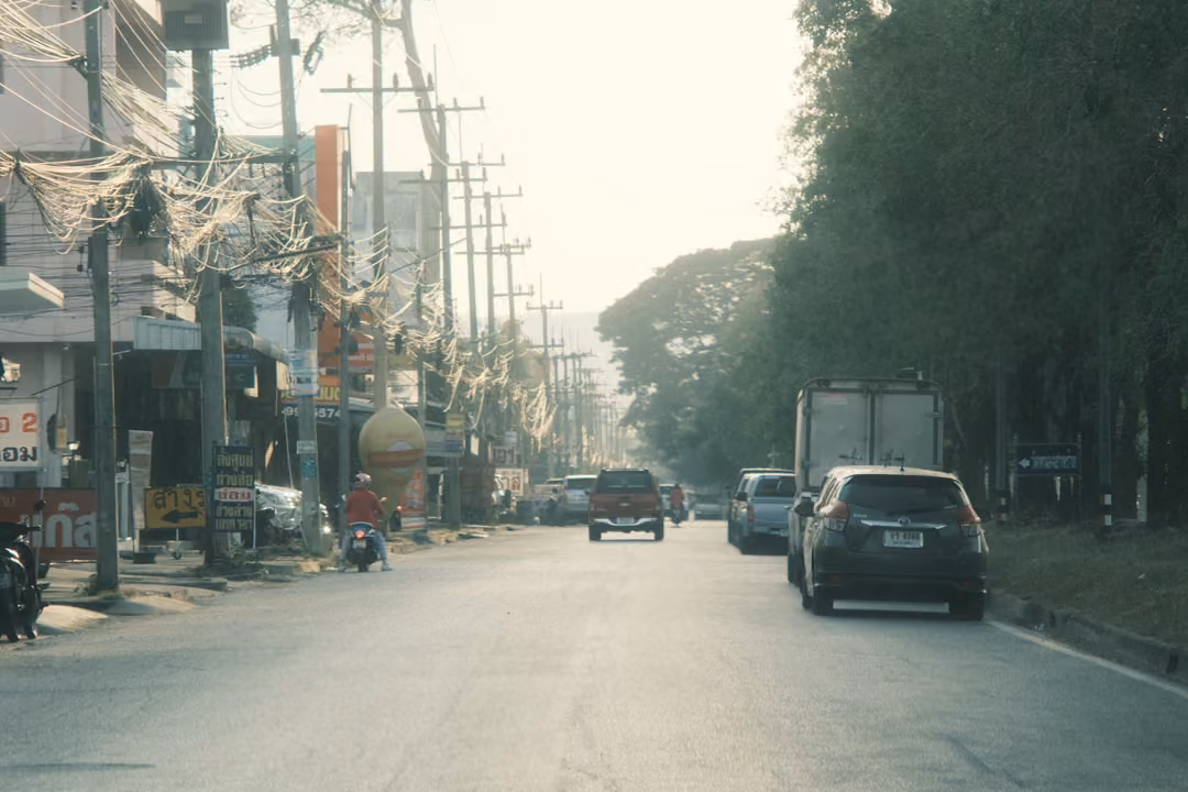 A long straight road in hazy light, cars and a truck ahead, tall trees on the right and looped cables on poles at left.