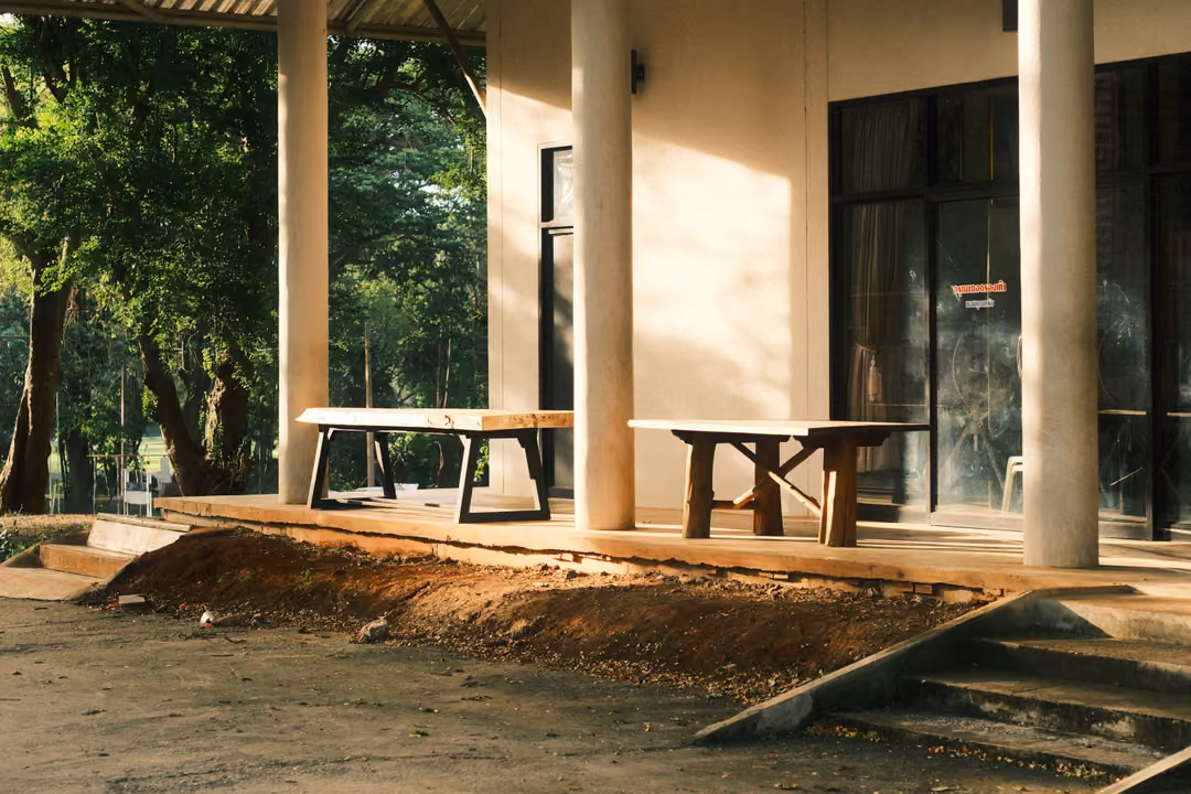 A veranda with pale columns and two wooden tables, steps leading down to red earth, lit by low golden sun.