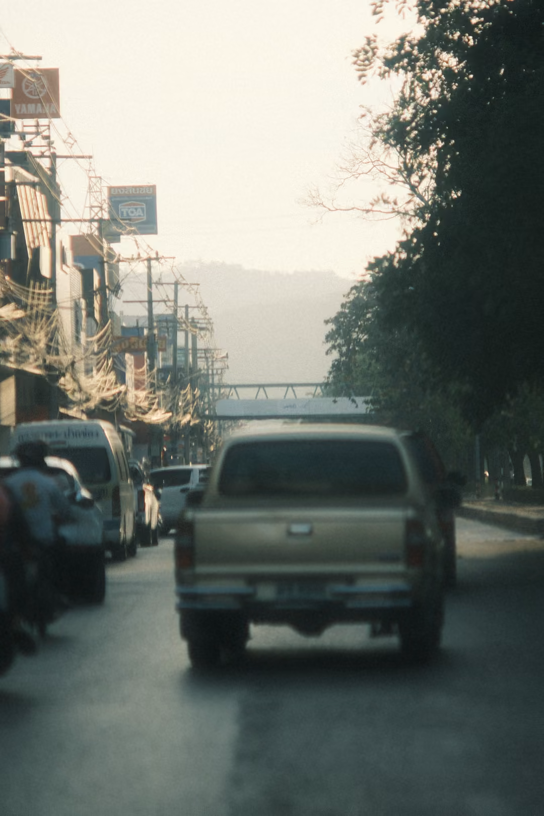 Traffic on a town road: a gold pickup, motorbike riders and a van, with a footbridge and hazy hills ahead.