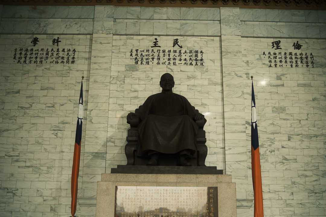 Darker view of the bronze statue between flags under a marble wall of carved text