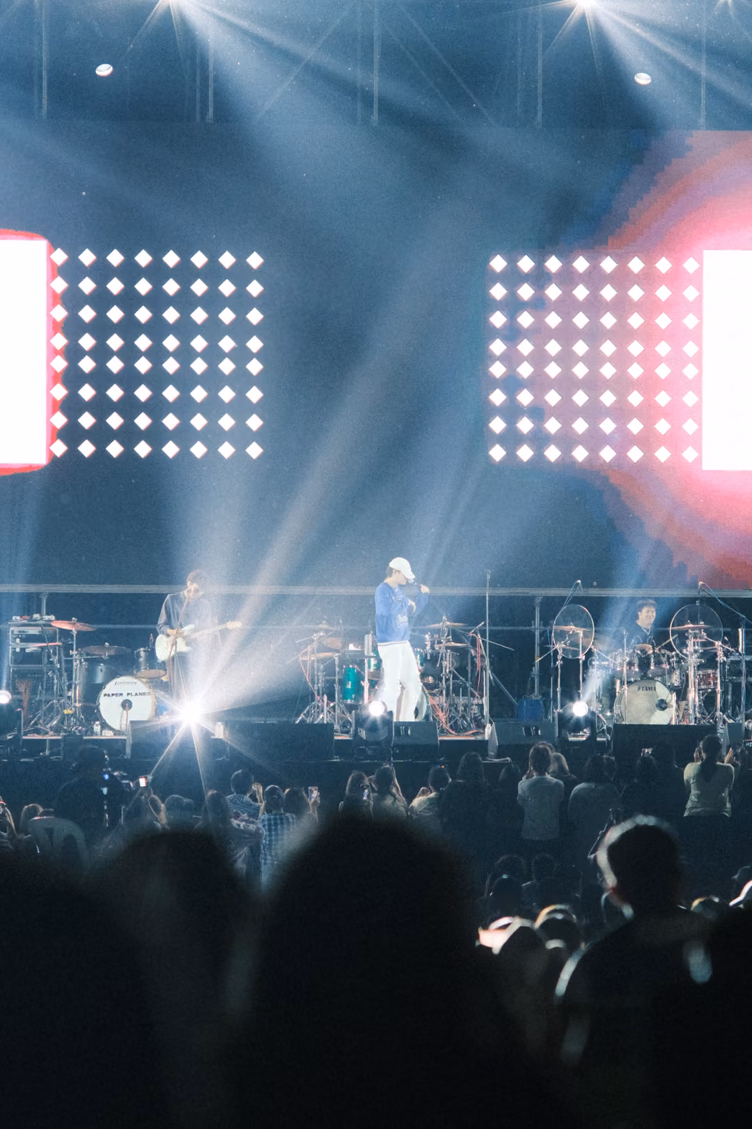 Vertical view of beams of light crossing over the stage, the singer at centre, pink glow on the screen at right.