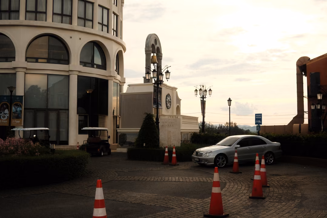 Forecourt with orange traffic cones, a parked silver car and golf carts by a round building and ornate lamps