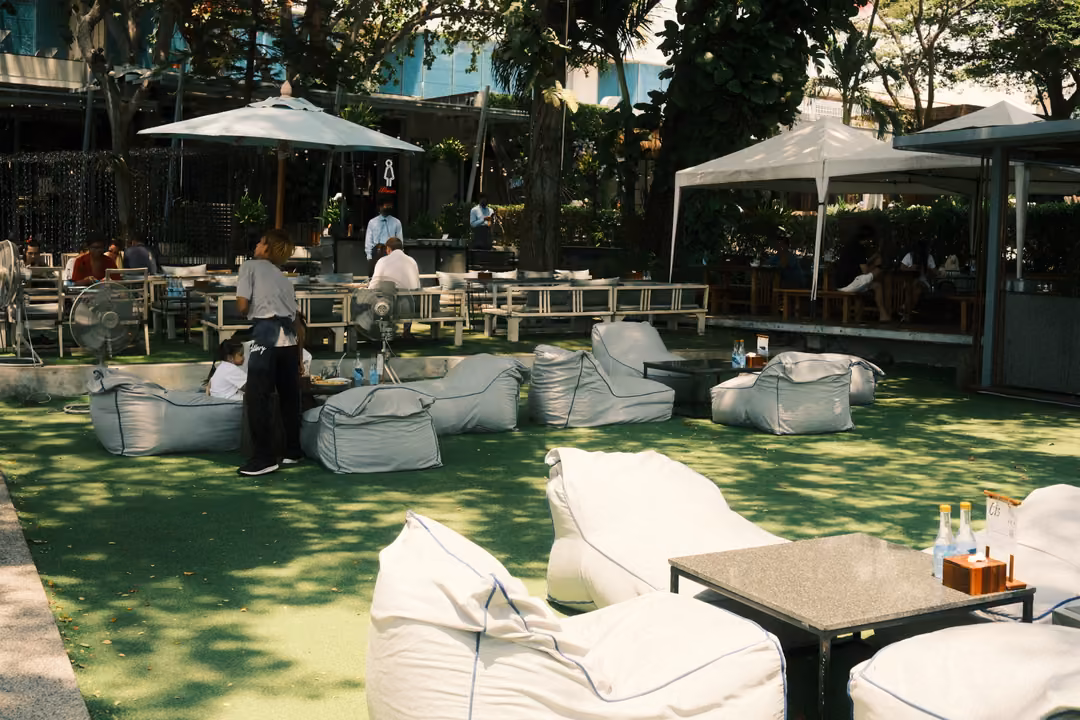 Lawn lounge with white beanbags, low tables, white umbrellas and people seated under shady trees