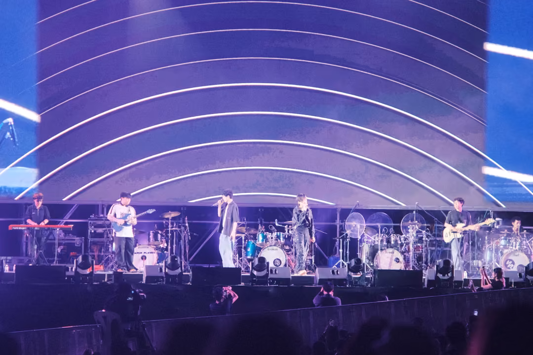 Band on stage in front of a giant arc-patterned violet screen, the singer at centre in pale trousers.