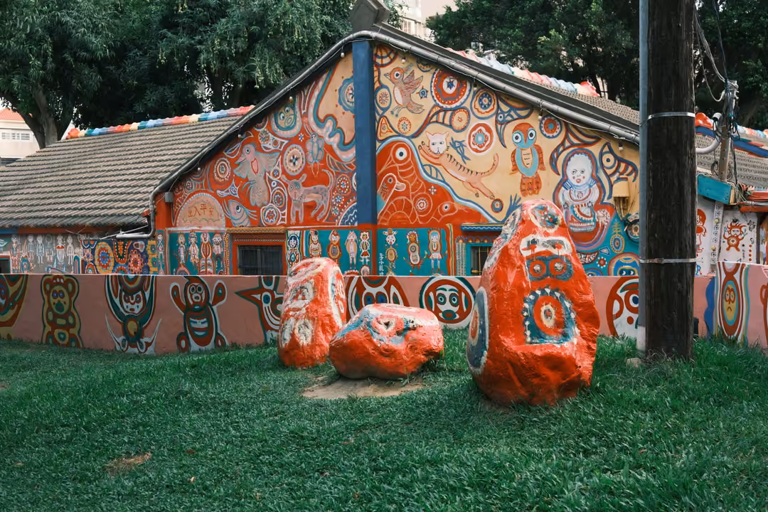 Painted house wall and gable with figures, three painted boulders on bright green grass