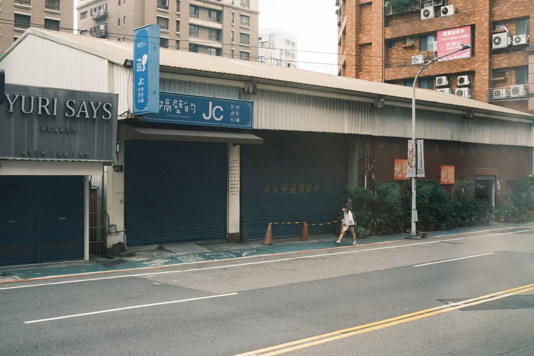 Street with a shop sign reading YURI SAYS, closed blue shutters, a pedestrian on the pavement, apartments behind