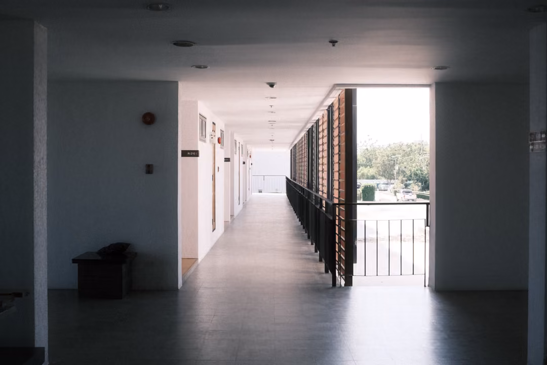 Long white hallway with doors on the left and slatted railings on the right, opening to bright daylight at the end
