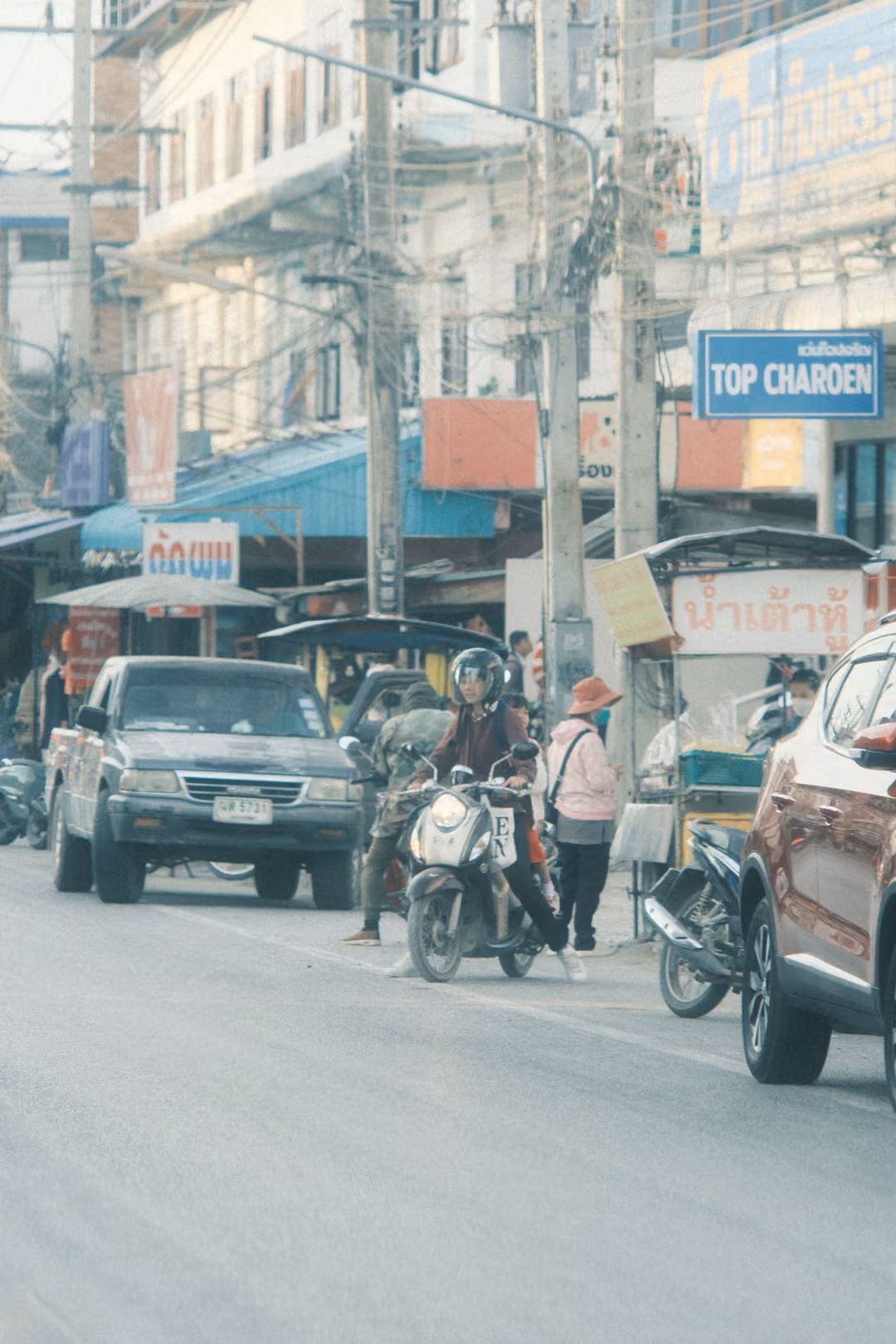 A town street with a pickup and a helmeted motorbike rider, shops and a blue TOP CHAROEN sign under tangled power lines.