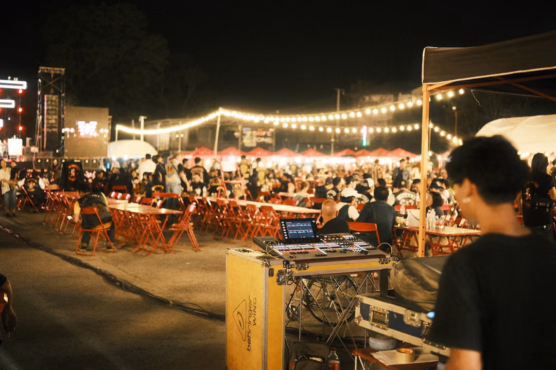 Man behind a sound mixing desk on a road case at a night festival, crowded tables and string lights behind