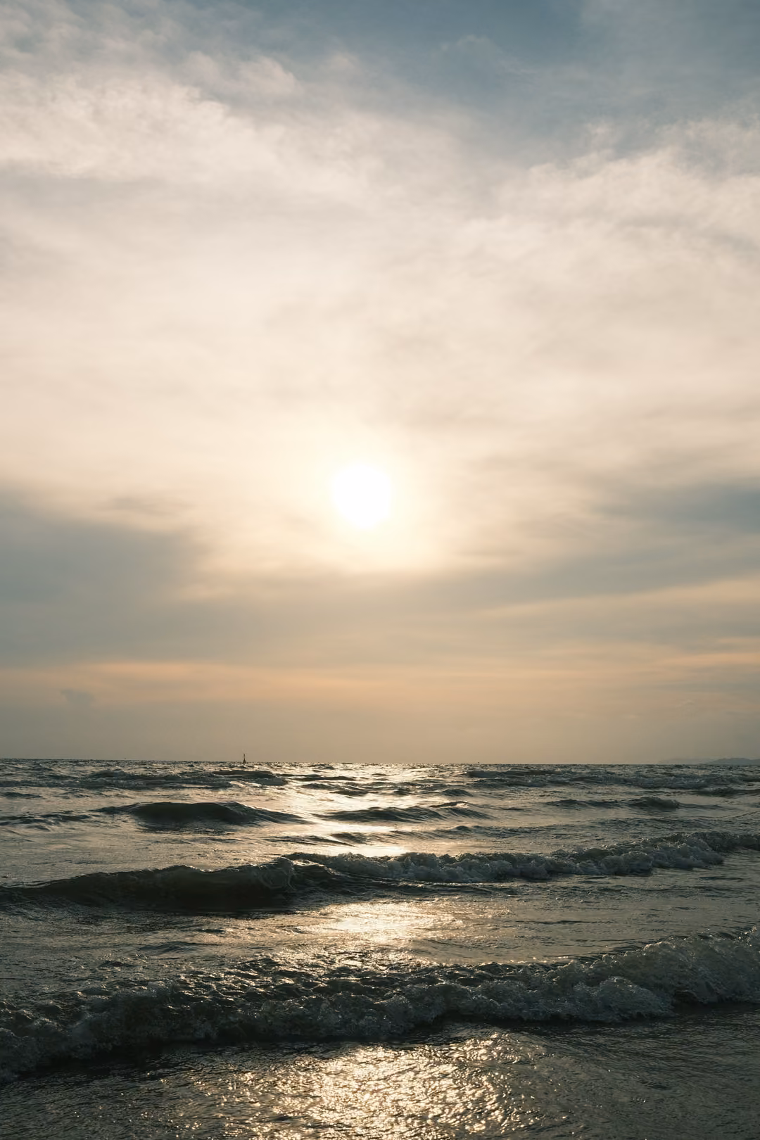 A low sun behind thin cloud above dark, foamy waves breaking on the shore