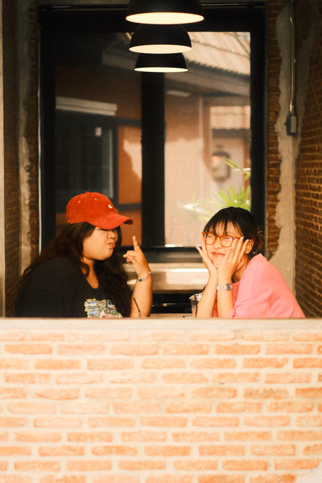 Two women at a window, one in a red cap pointing a finger, the other in pink with face cupped in her hands
