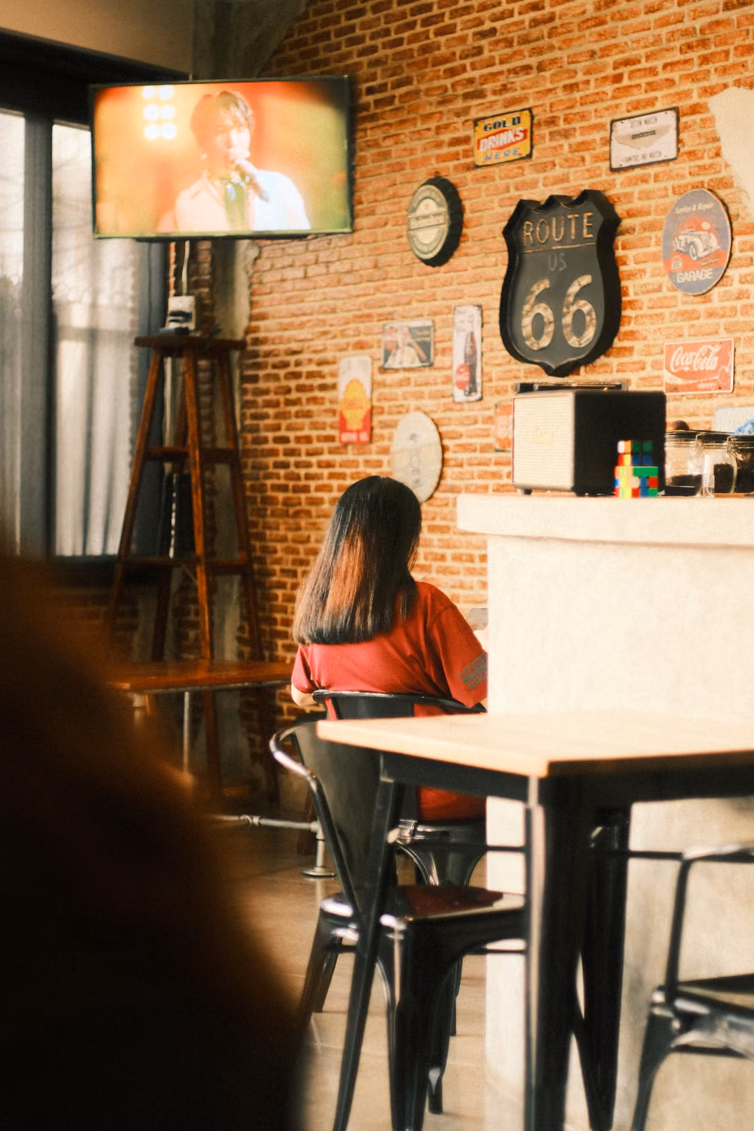 A person with long hair sits at a cafe table seen from behind, a TV, clocks and a Route 66 sign on the brick wall