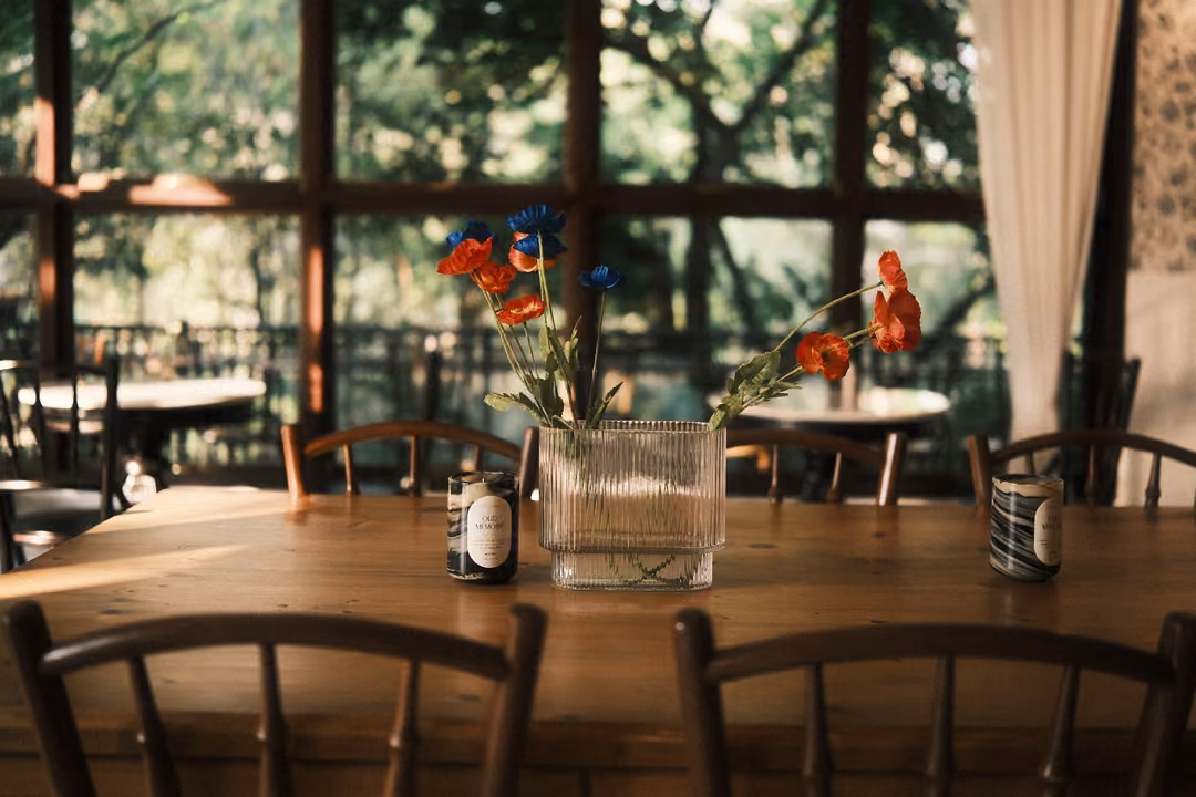 Long wooden table with a ribbed glass vase of red and blue flowers and two cans, windows onto trees behind