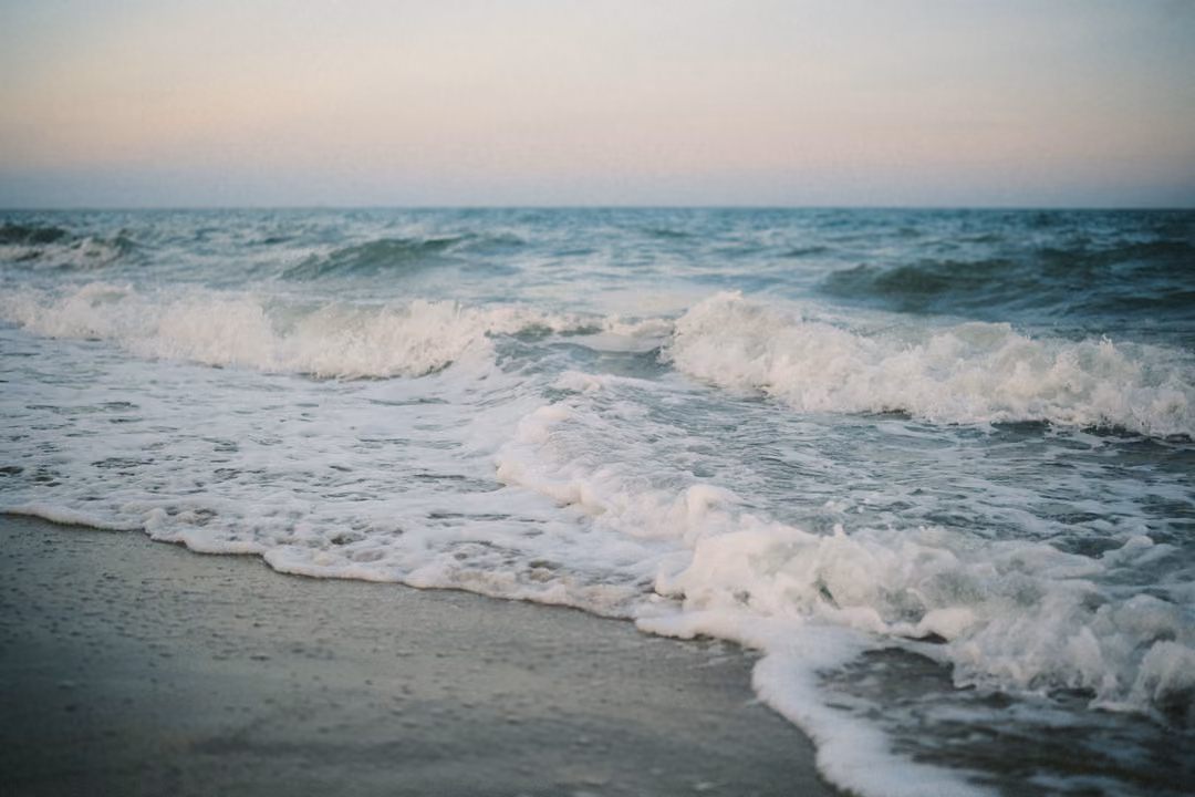 Waves breaking white on a shore at dusk, the sea teal and the sky pale pink at the horizon