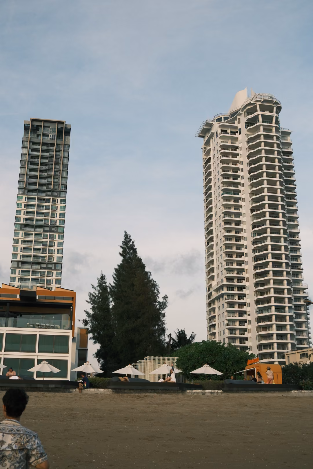Two tall residential towers and a dark tree rising behind beach umbrellas, a person in the foreground