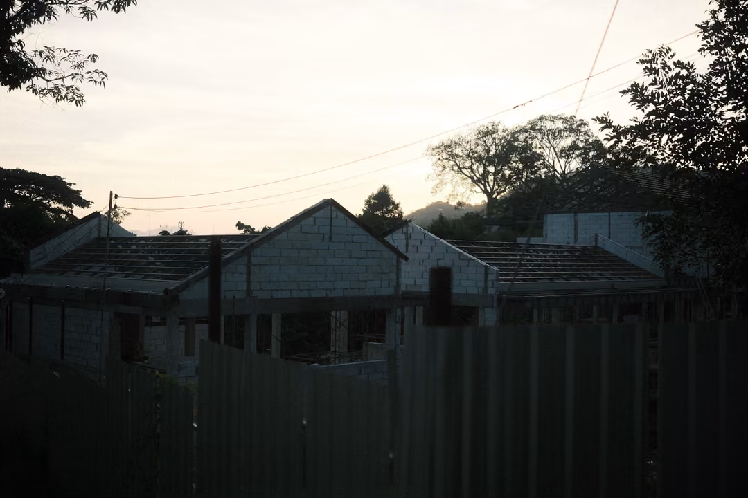 Unfinished houses behind a corrugated fence in dim evening light, trees and wires against the sky