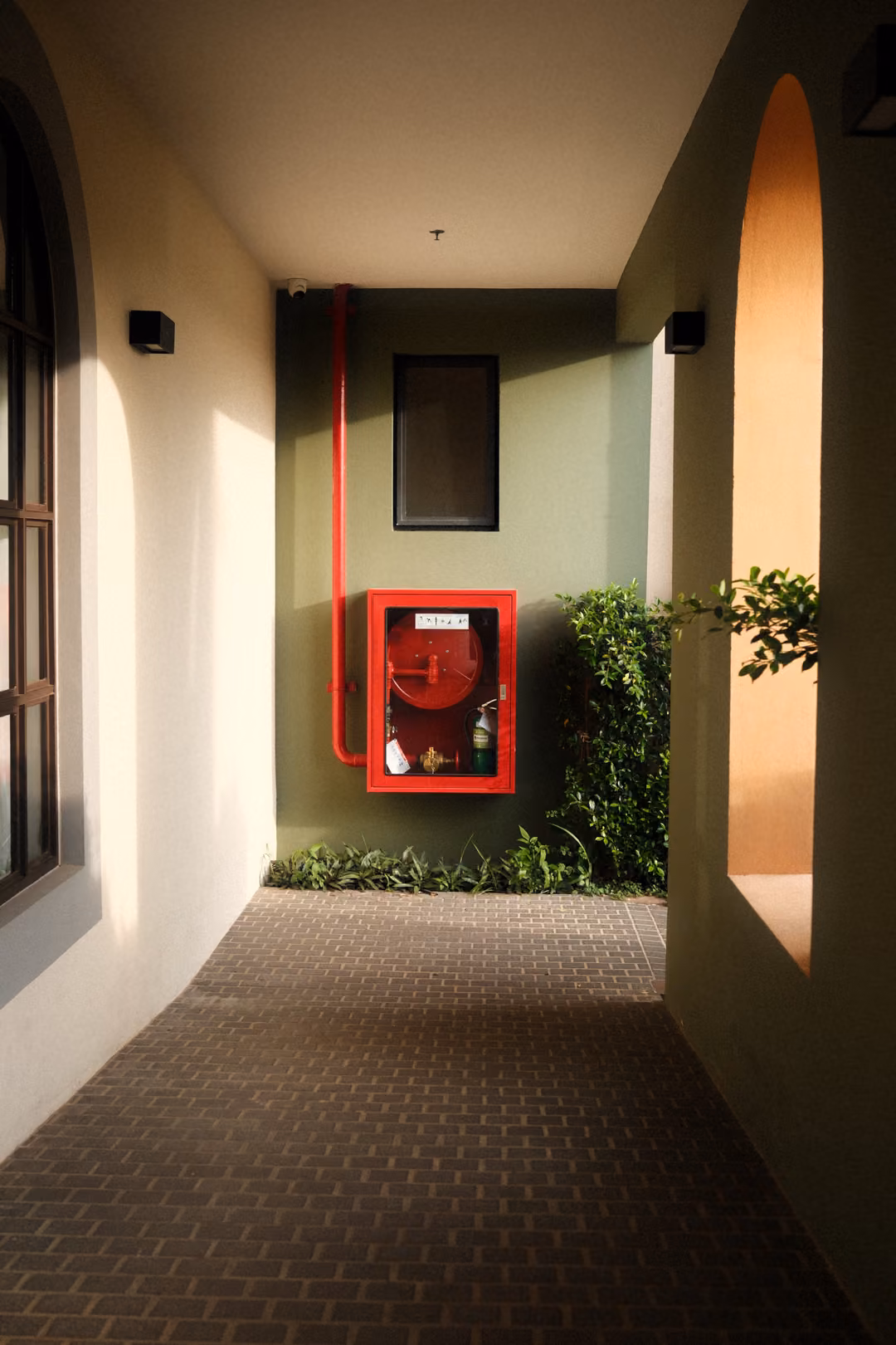 Arcade corridor with brick paving leading to a green wall with a red fire hose cabinet and plants
