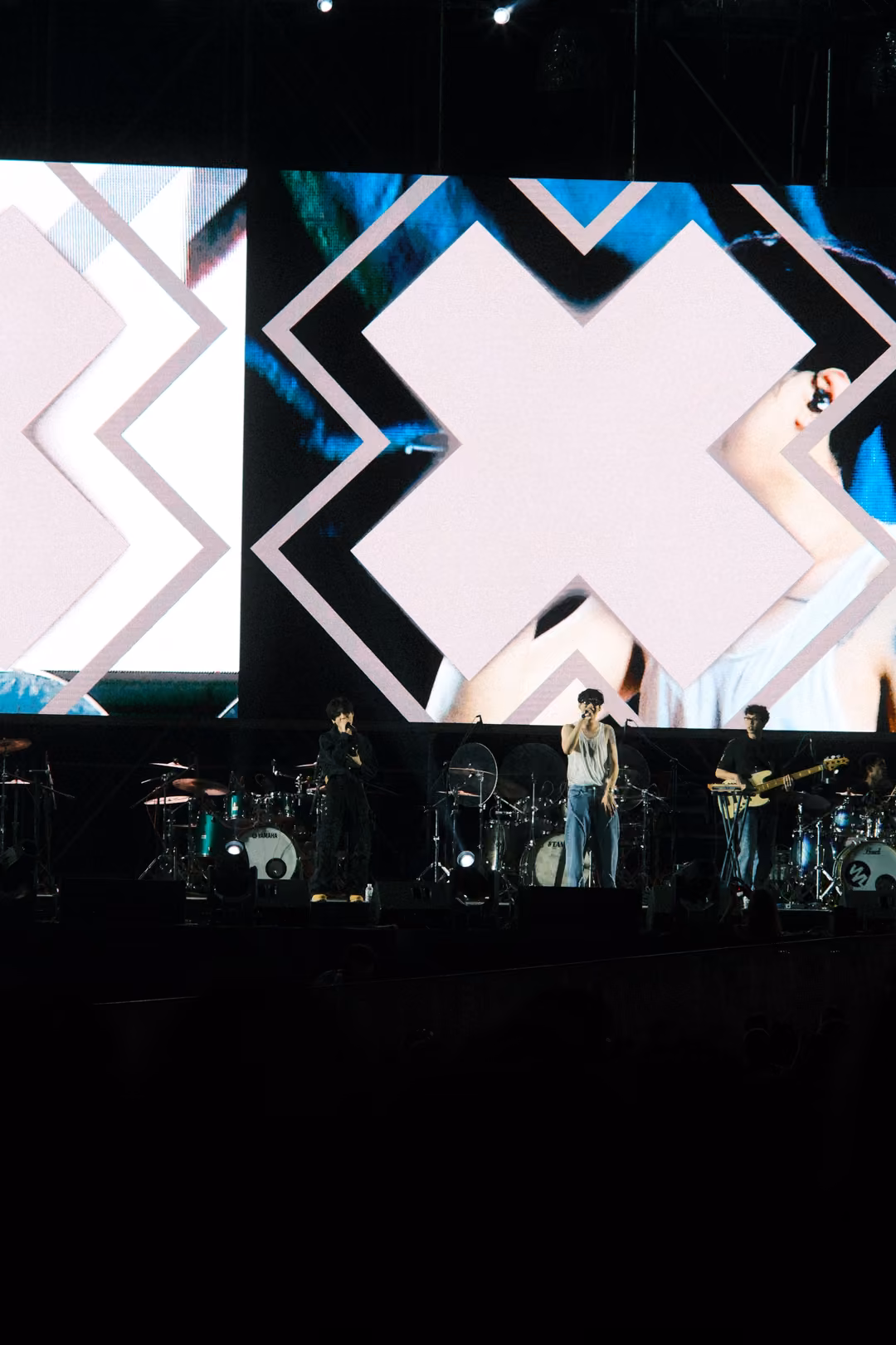 Screen with a giant pale X shape over a close-up of a singer with a microphone, band on stage beneath.
