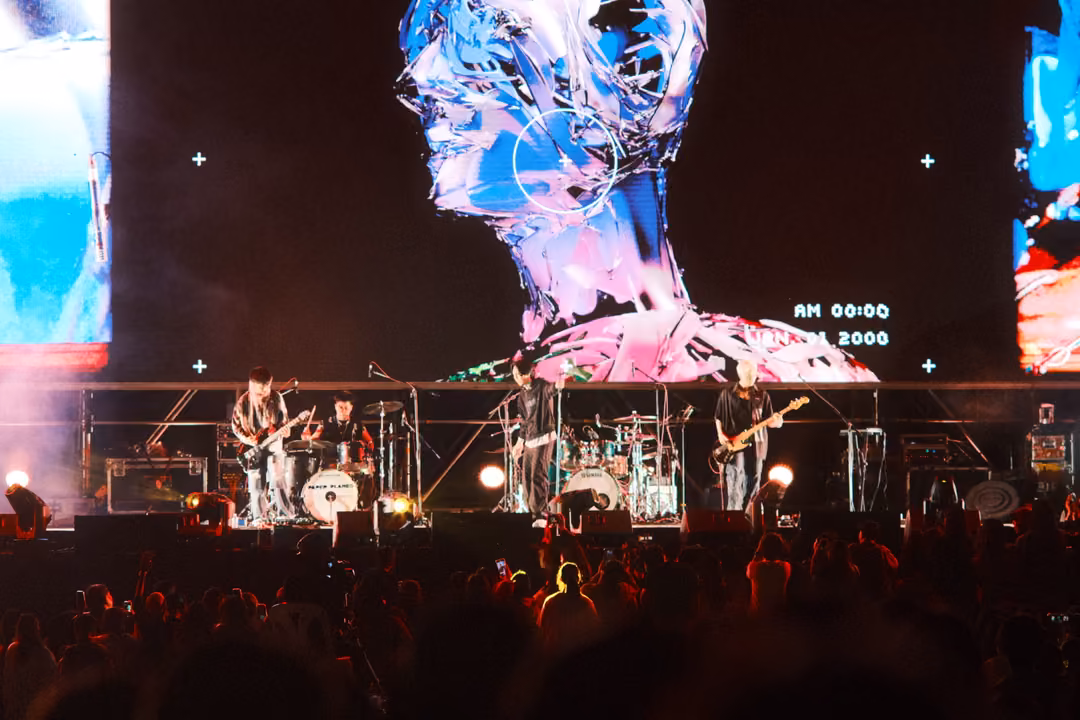 Same stage a moment later, the chrome head on the screen tinted pink and blue above the band and dark crowd