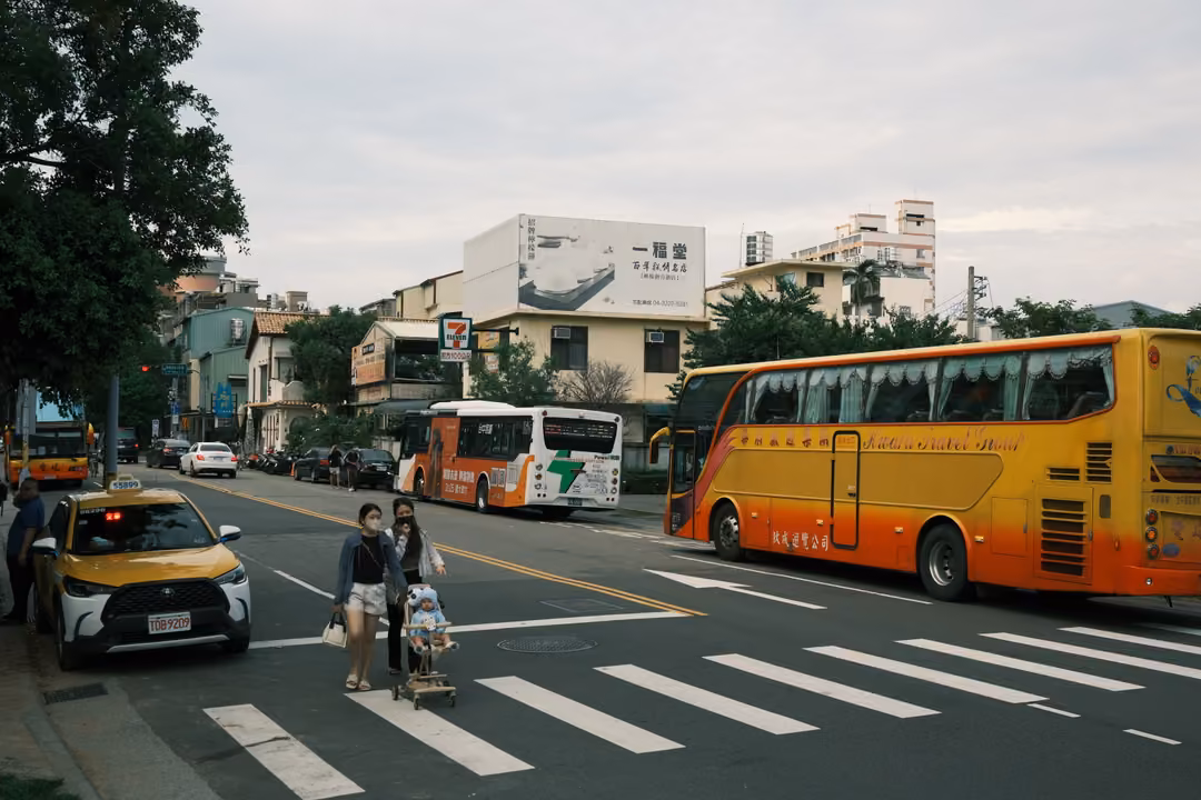 Two people with a pushchair crossing a city street on a crosswalk, a yellow coach bus and taxi nearby
