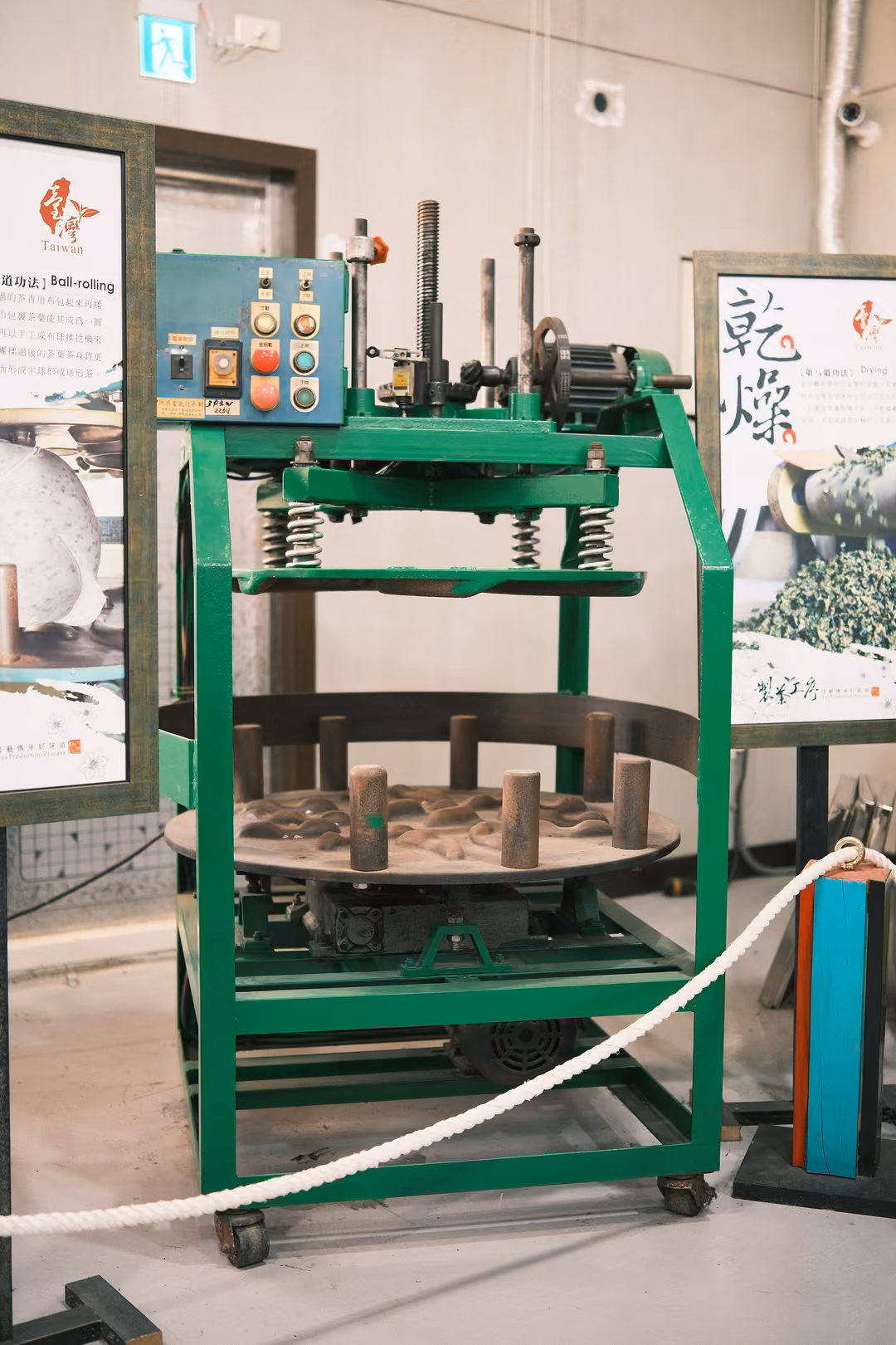 Same green tea-rolling machine seen slightly wider, rope barrier curving in front, posters on both sides