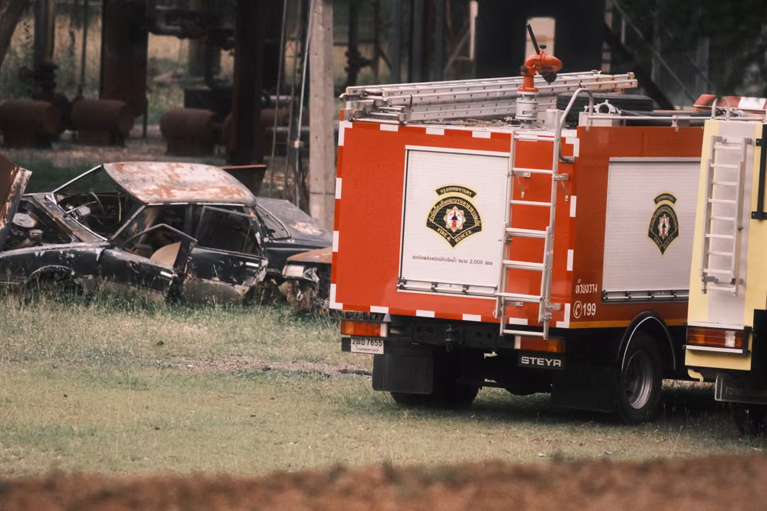 Rear of a red fire truck with ladders and emblem panels, an old wrecked car parked in the grass beside it