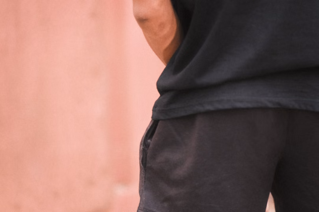 Close crop of a person's side in a black T-shirt and dark shorts against a pink wall, arm at their side
