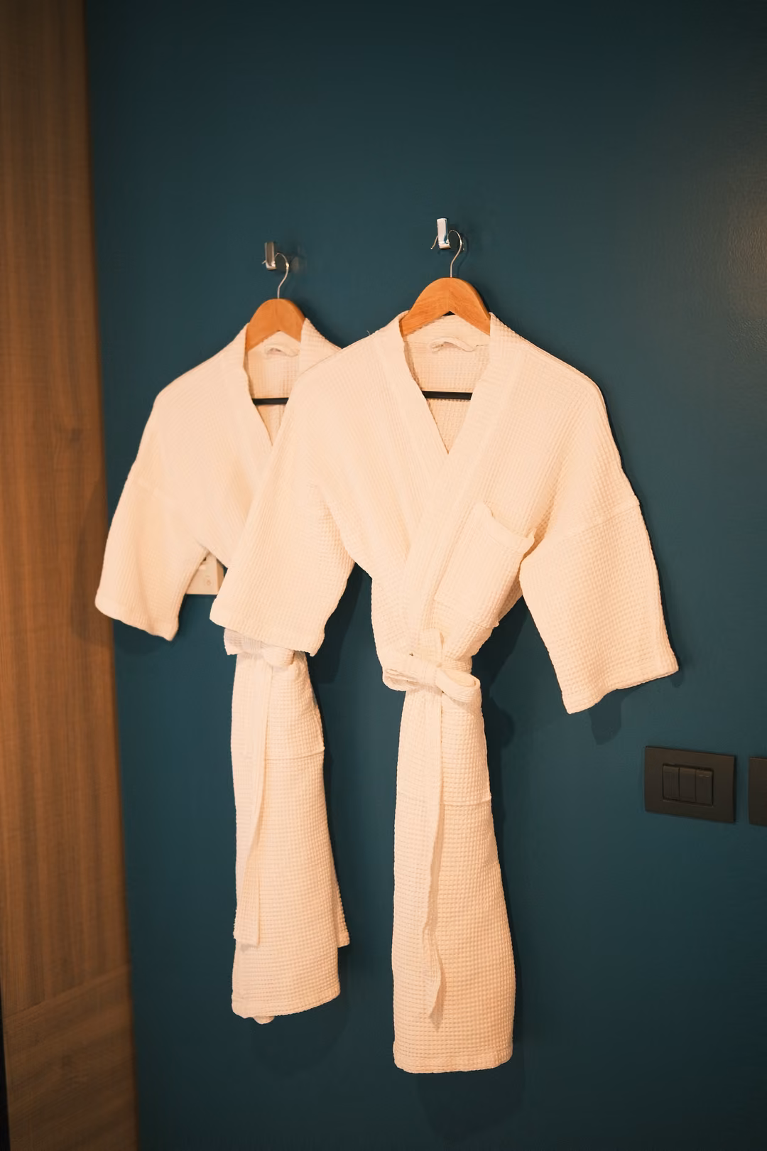 Two white bathrobes hanging on hangers against a dark teal wall