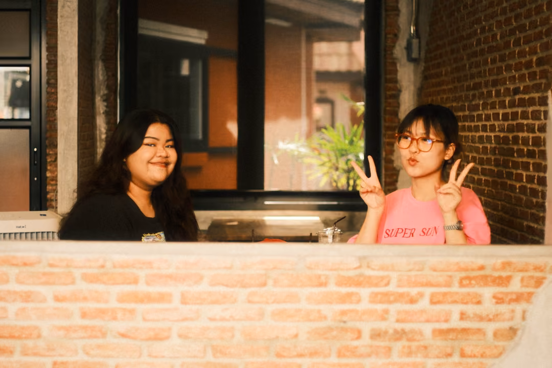 Two women behind a brick wall, one in pink raising two peace signs, one in black smiling