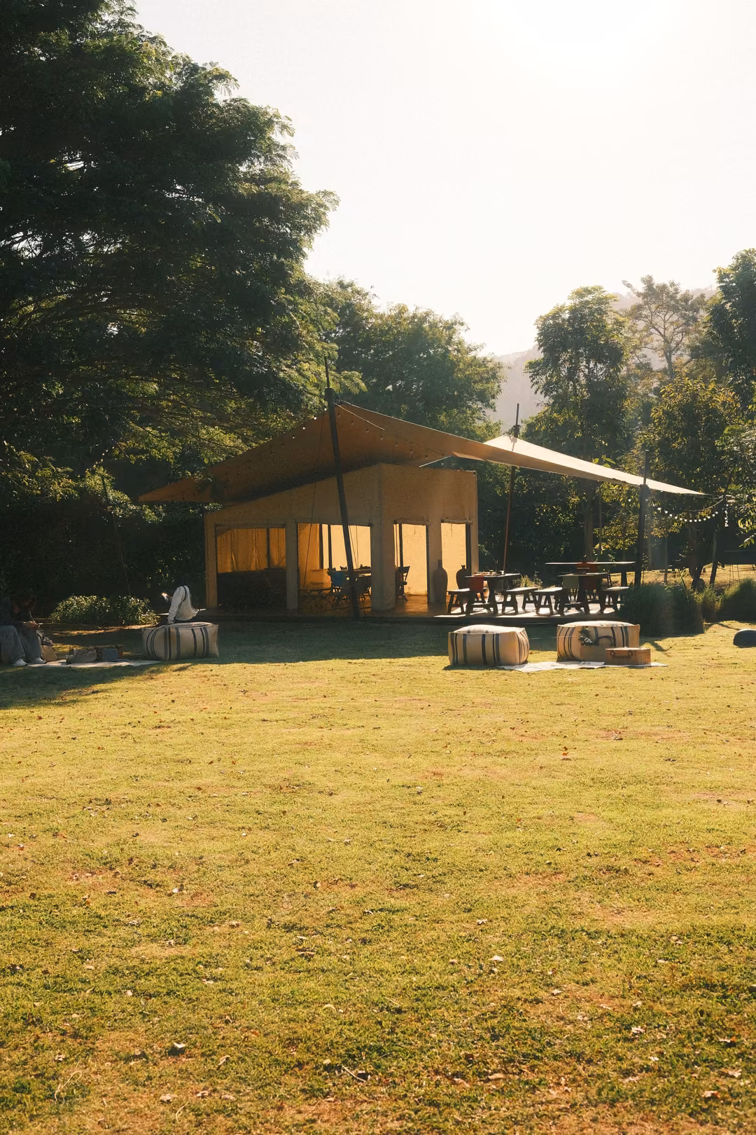 Canvas-roofed pavilion on a sunlit lawn, striped poufs set out on the grass, tall trees behind