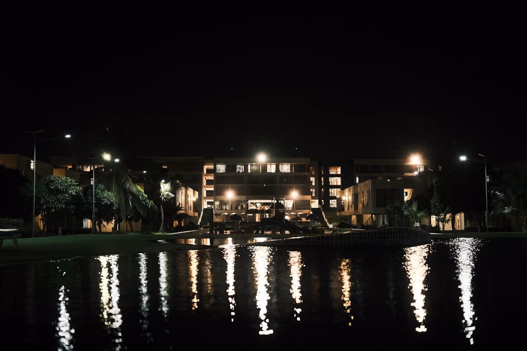 Resort buildings and street lamps reflected in a dark pond at night, a lawn and palms along the edge