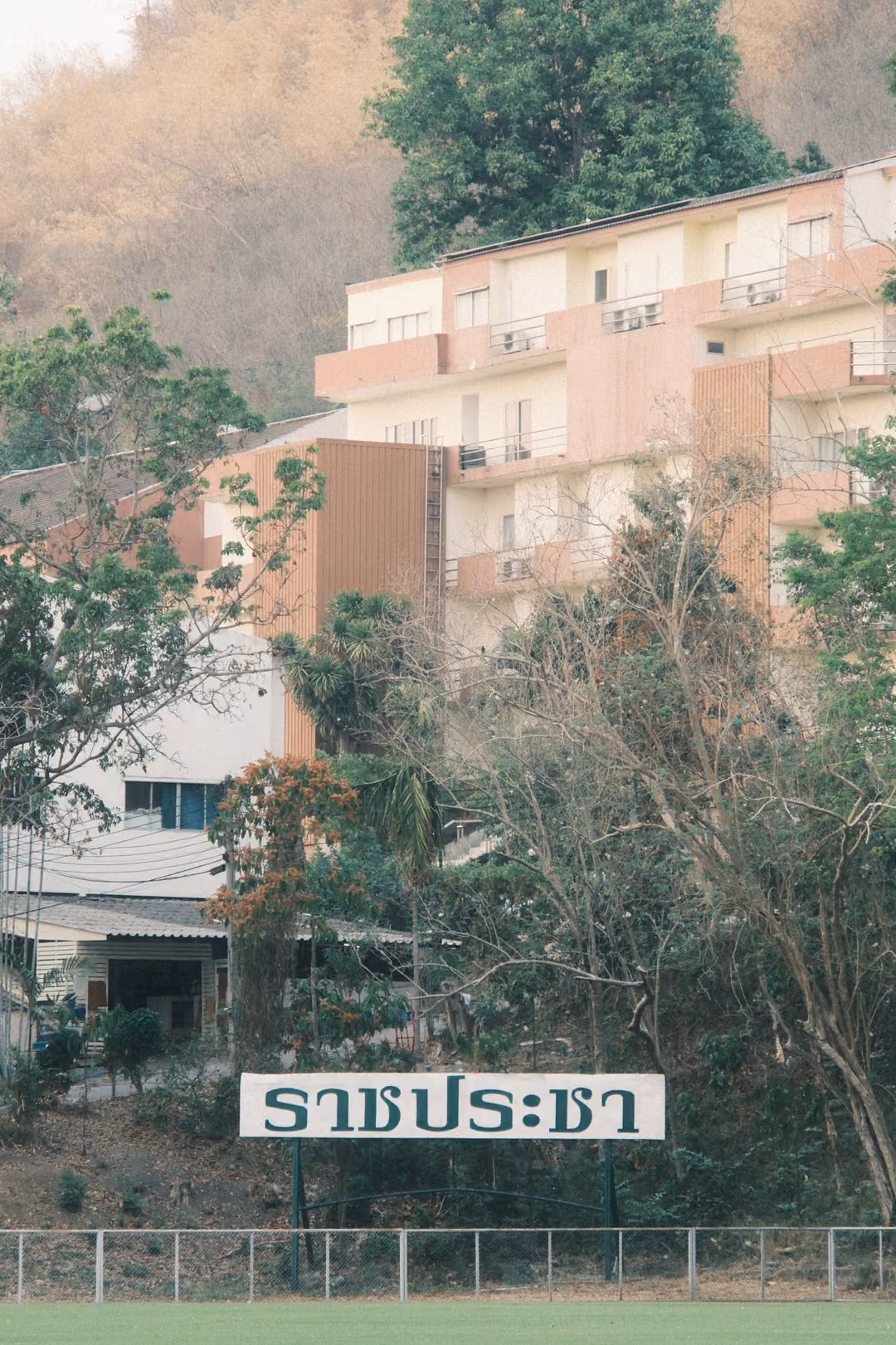 Vertical view of a peach and white hillside building behind trees, a white sign with Thai lettering below