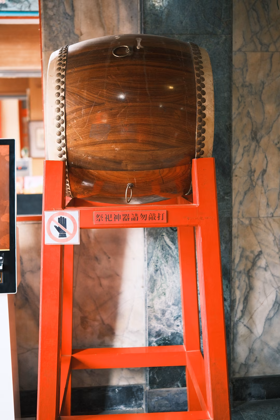 Large wooden drum with a glossy grain mounted on a tall red stand against a marble wall, a no-touch sign below