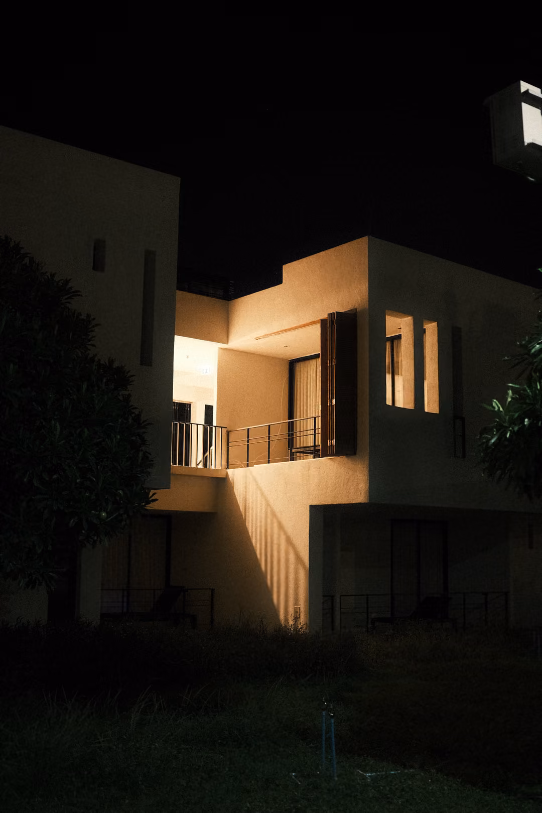 A white modern two-storey villa lit warmly at night against a black sky, a tree to the left