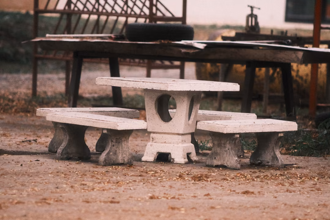 Concrete picnic table and benches on dirt ground, an old wooden table and metal rack behind