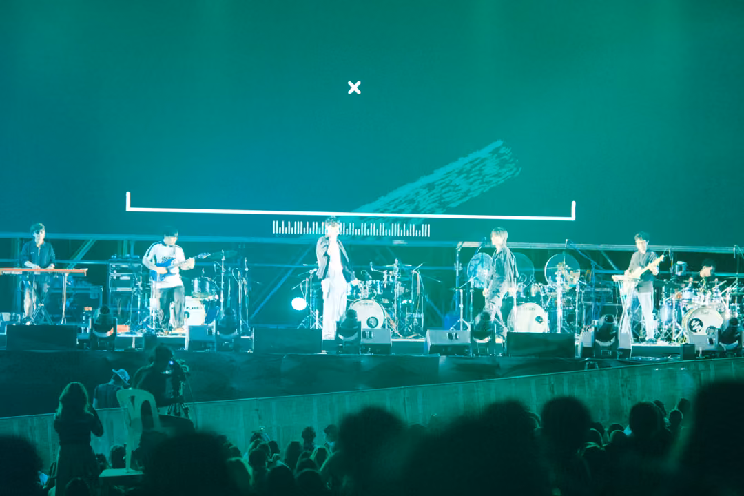 Band on stage bathed in green light, a white bracket shape and small cross on the screen behind them.