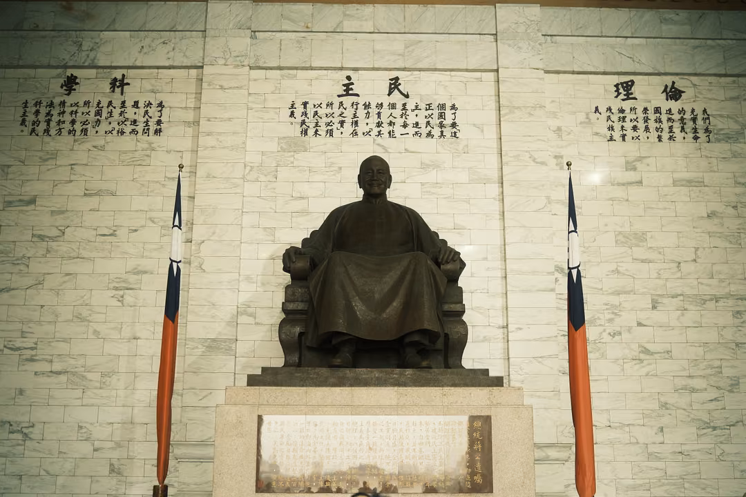 Bronze seated statue centred between two flags below marble wall inscriptions, lit warm from above