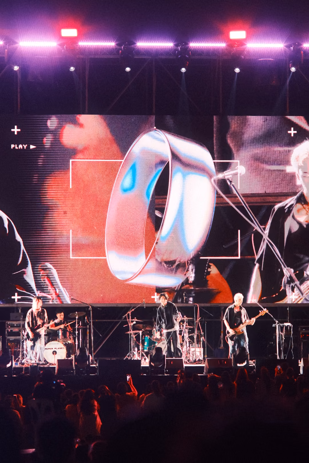 Vertical view of a glossy ring on screen with a PLAY label, pink lights above, three musicians performing below.