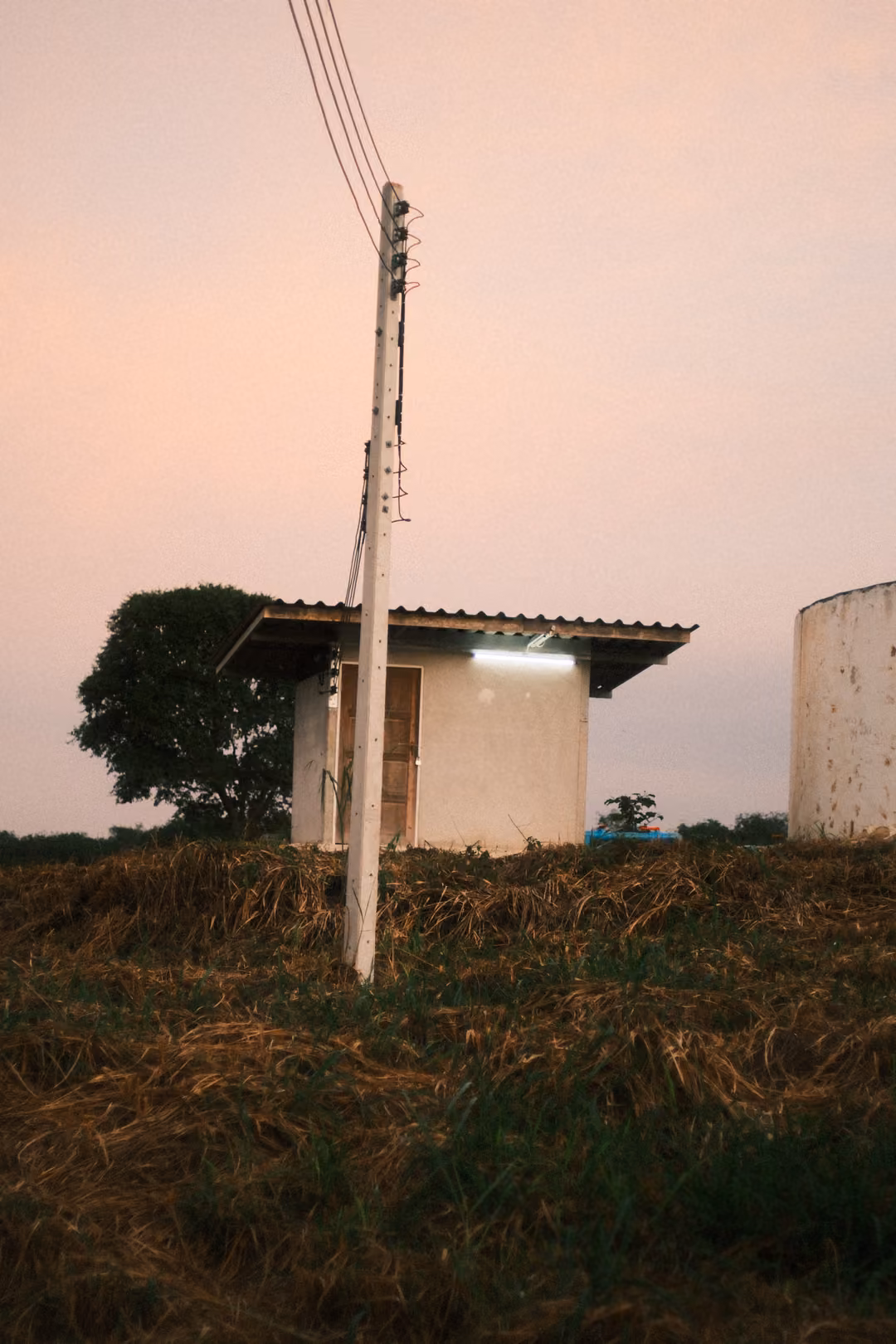 Small white building with a lit fluorescent light, a power pole and a water tank under a pink sky