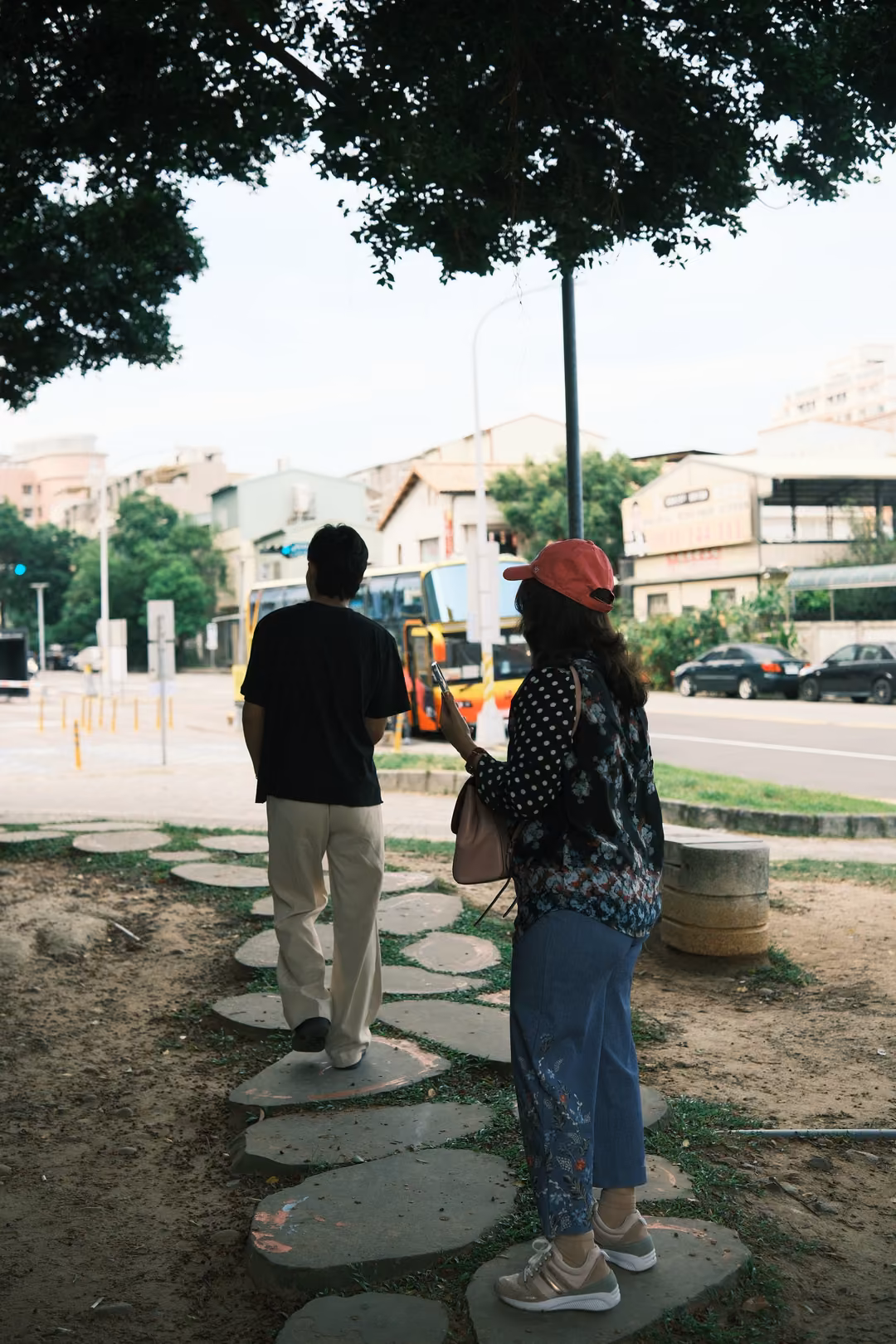 Man and woman seen from behind on a stepping-stone path under a tree, a yellow bus and street beyond