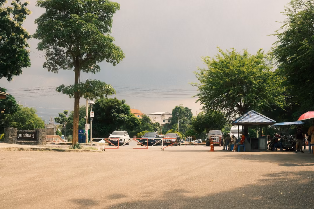 Wide parking area under an overcast sky, tall trees, cars and a small stall with umbrellas at the edge
