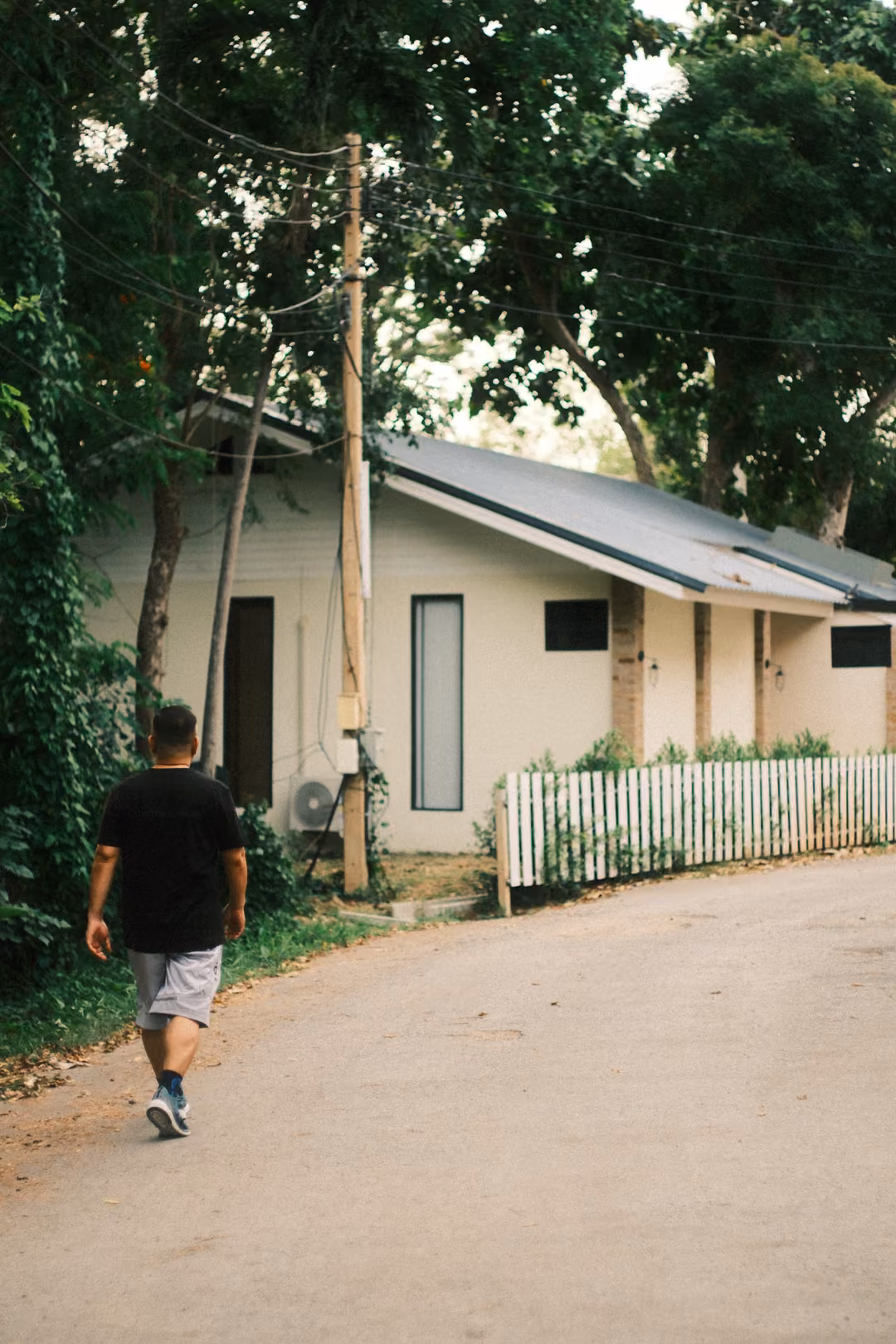 A figure in a black T-shirt and grey shorts walks along a road past a white cottage with a picket fence.